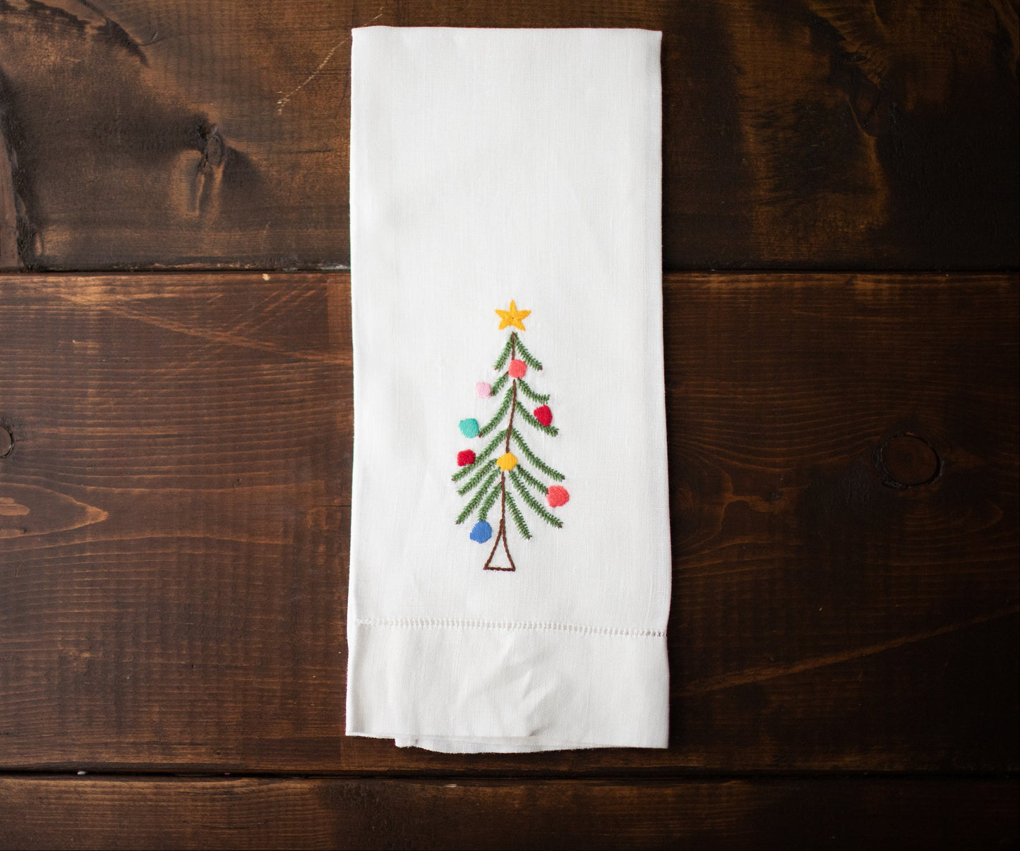 A white linen tea towel with a hand-embroidered Christmas tree design, laid out on a wooden surface.