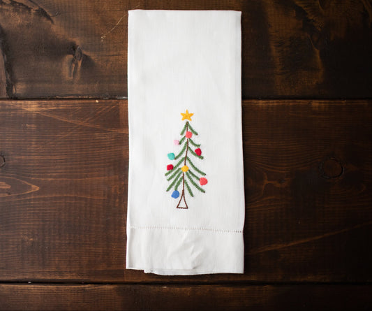 A white linen tea towel with a hand-embroidered Christmas tree design, laid out on a wooden surface.