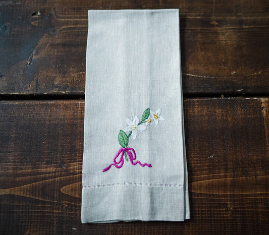 Folded flax linen tea towels with hand embroidered floral designs and a bow motif, displayed on a wooden surface.